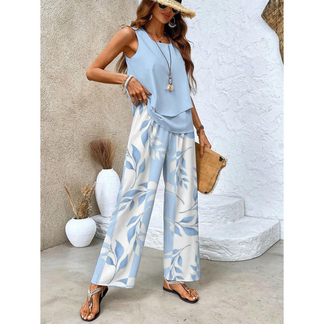 Femme en tenue estivale chic, portant un ensemble bleu à motifs floraux, sandales dorées, chapeau de paille, posant près de vases décoratifs blancs.