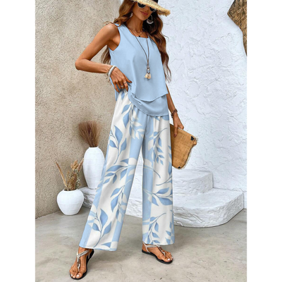 Femme en tenue estivale chic, portant un ensemble bleu à motifs floraux, sandales dorées, chapeau de paille, posant près de vases décoratifs blancs.