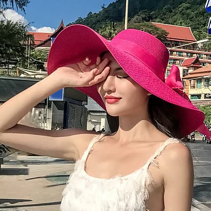 Femme élégante portant un chapeau rose vif, robe blanche, souriant sous le soleil, paysage urbain en arrière-plan, journée d'été.