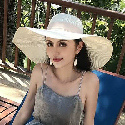 Jeune femme portant un chapeau de soleil blanc, robe grise, assise sur une chaise bleue en extérieur, entourée de verdure tropicale.