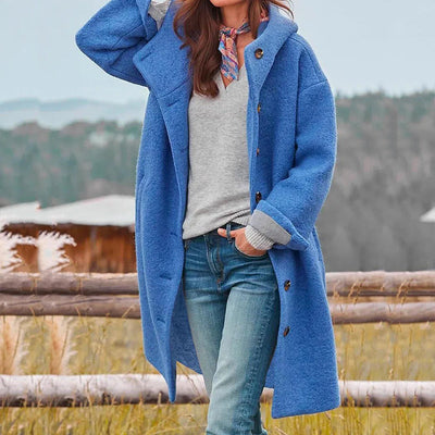 Femme portant un manteau bleu en laine, écharpe colorée, pull gris et jeans, posant devant une clôture en bois avec paysage naturel en arrière-plan.