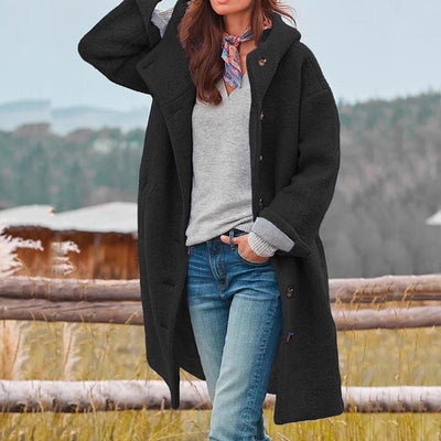 Femme portant un manteau noir élégant, pull gris et jean, posant devant un paysage naturel. Mode automne-hiver, style décontracté chic.