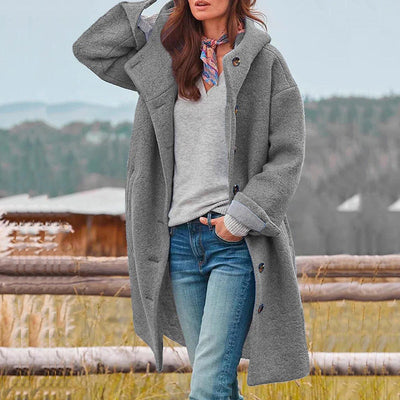 Femme portant un manteau gris en laine, chemise grise, foulard coloré, posant devant un paysage rural avec clôture en bois et collines.
