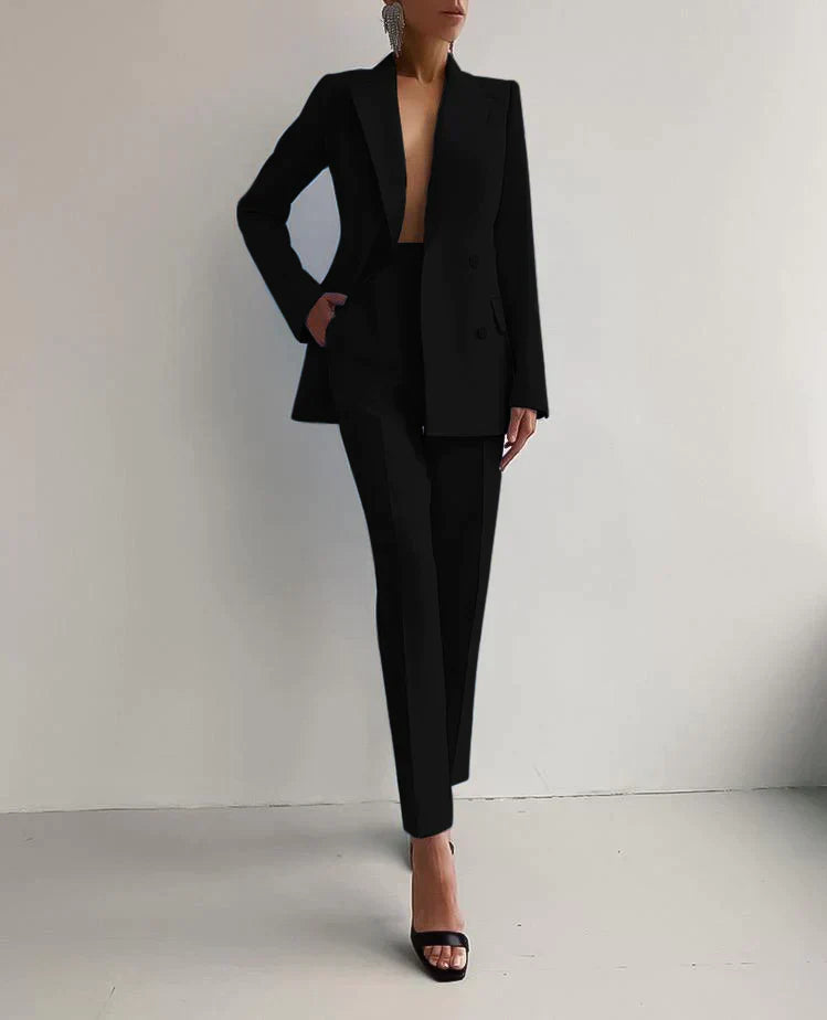 Femme en tailleur noir élégant, veste et pantalon, posant contre un mur blanc. Mode chic, tenue professionnelle, style minimaliste.