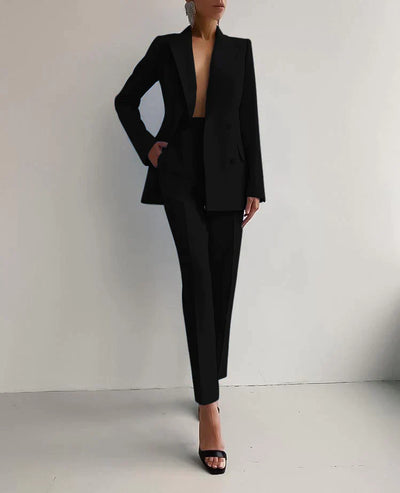 Femme en tailleur noir élégant, veste et pantalon, posant contre un mur blanc. Mode chic, tenue professionnelle, style minimaliste.