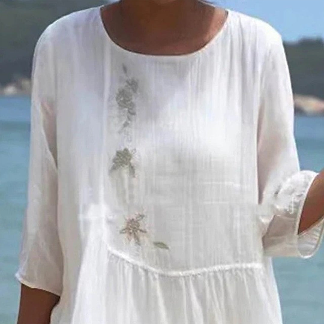 Robe blanche en lin avec broderie florale, manches trois-quarts, portée en bord de mer. Mode estivale élégante, tenue légère pour plage.