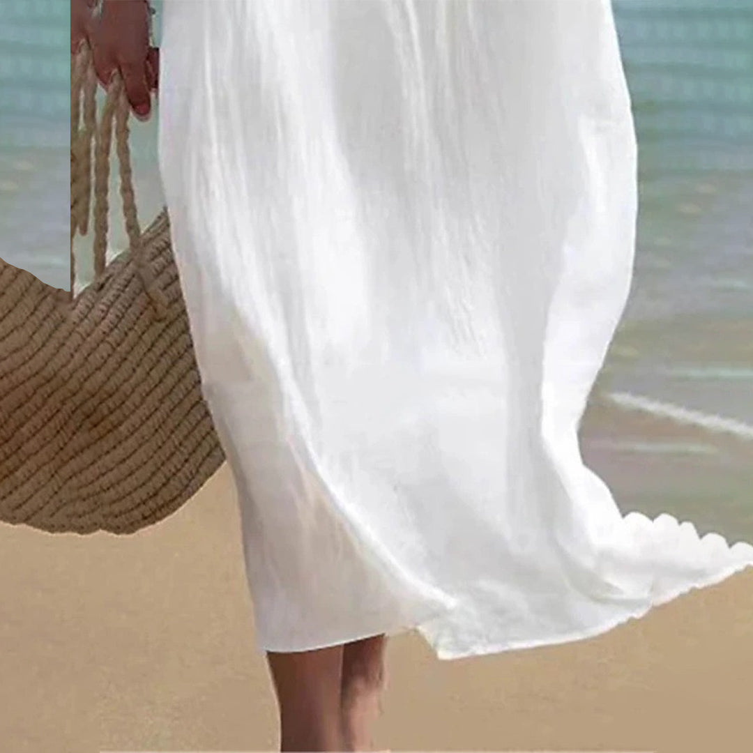 Robe blanche fluide sur plage, femme tenant sac en paille, bord de mer, mode estivale, style décontracté, vacances, élégance simple.