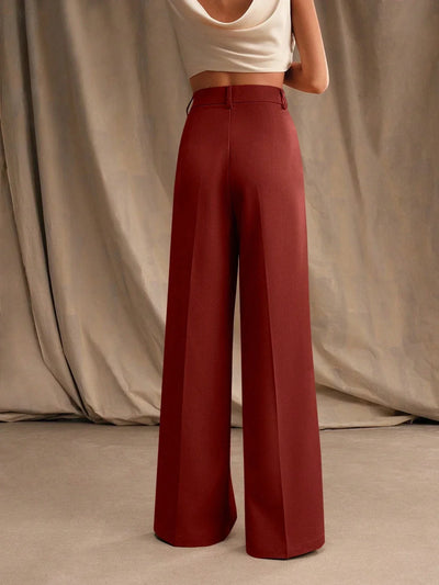 Femme portant un pantalon large rouge bordeaux et un haut blanc sans manches, posant devant un rideau beige. Mode élégante et tendance.