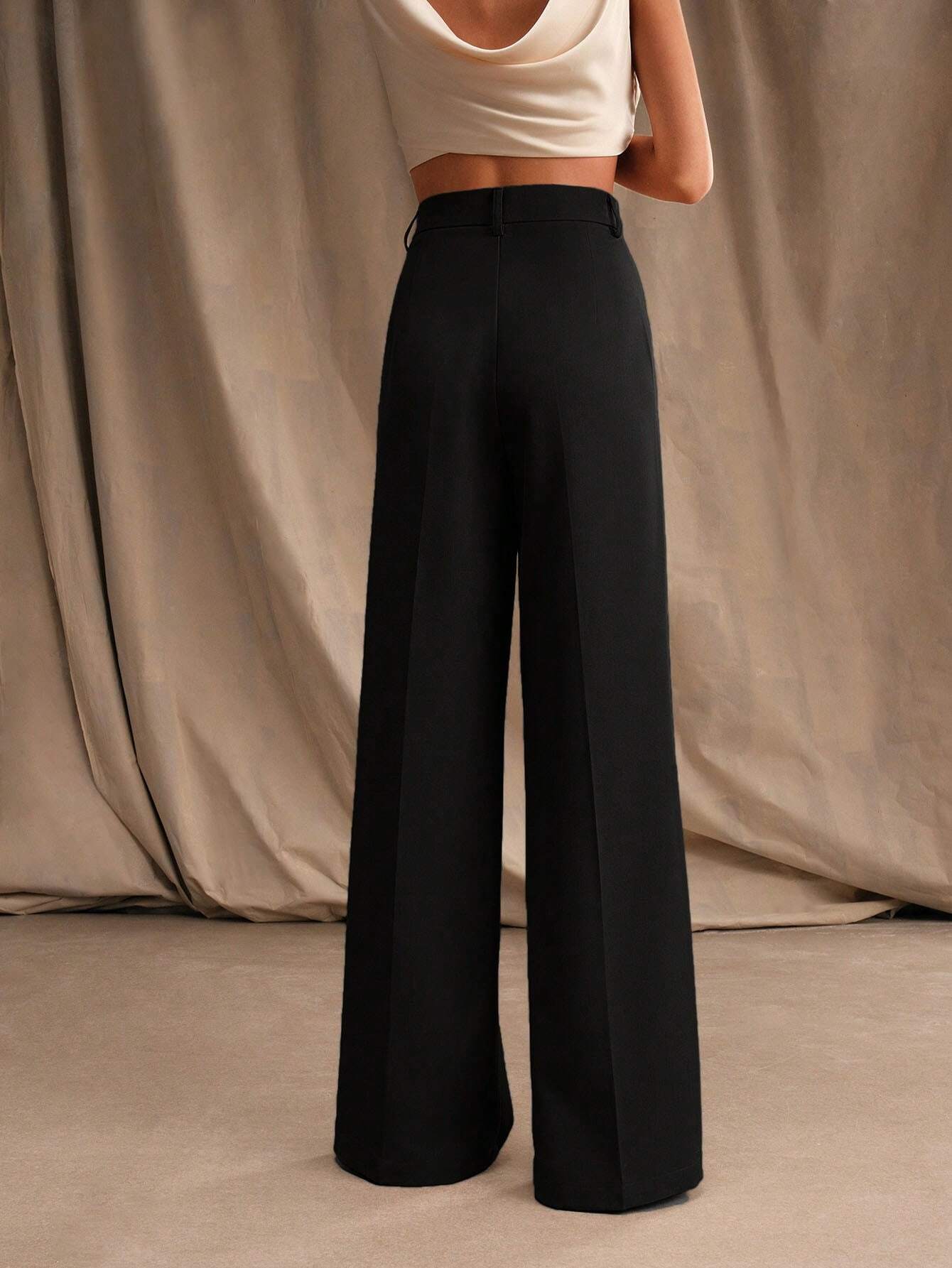 Femme portant un pantalon large noir taille haute, associé à un haut blanc sans manches, posant devant un rideau beige. Mode élégante et minimaliste.