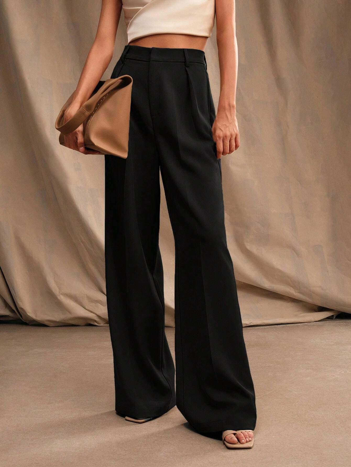 Femme portant un pantalon noir large et un haut blanc, tenant un sac à main beige. Mode élégante, style minimaliste, tenue chic.