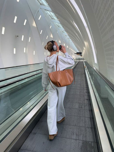 Femme en survêtement gris et casque audio, portant un grand sac marron, marche sur un tapis roulant dans un aéroport moderne et lumineux.