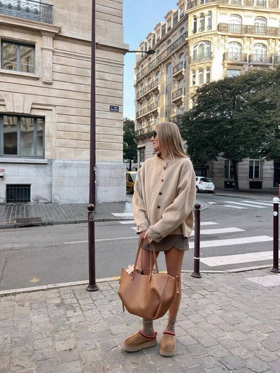 Femme en manteau beige et bottes UGG, tenant un sac en cuir marron, traverse une rue parisienne bordée de bâtiments haussmanniens.