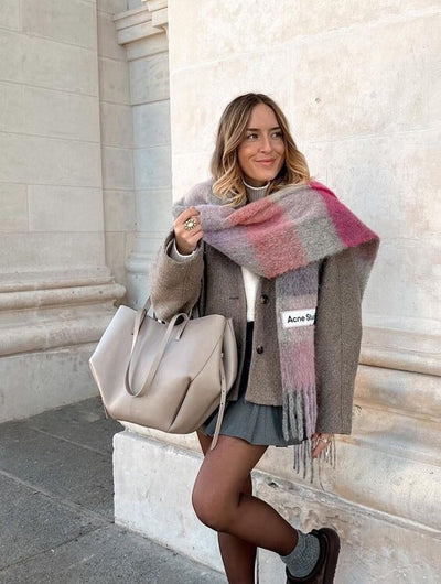 Jeune femme souriante portant une écharpe à carreaux rose et grise, manteau beige, jupe grise, sac à main beige, posant devant un mur en pierre.