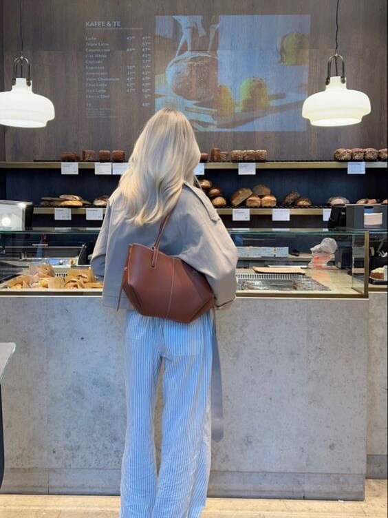 Femme blonde devant une vitrine de boulangerie, portant un sac marron, avec des pâtisseries en arrière-plan. Éclairage moderne, ambiance café.