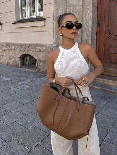 Femme élégante portant un débardeur blanc et un grand sac à main marron, lunettes de soleil noires, posant devant un bâtiment en pierre.