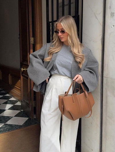 Femme élégante en manteau gris et pantalon blanc, portant un sac à main marron. Mode automne chic, lunettes de soleil, style urbain moderne.