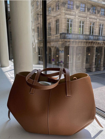 Sac à main en cuir marron sur une table près d'une fenêtre, avec vue sur un bâtiment historique parisien. Accessoire mode élégant et tendance.