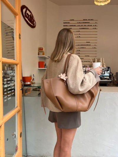 Femme de dos avec sac à main marron et gobelet café, devant un comptoir de café moderne. Menu visible en arrière-plan. Mode décontractée.
