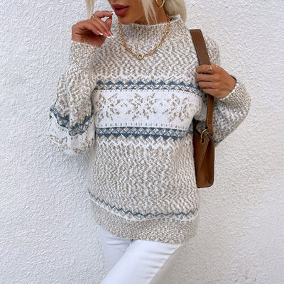 Femme portant un pull tricoté beige à motifs bleus et blancs, avec un sac marron, posant contre un mur blanc. Mode automne-hiver élégante.