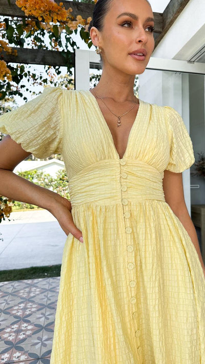 Robe jaune à manches courtes avec boutons, portée par une femme souriante en extérieur, fond floral et carrelage décoratif. Mode estivale élégante.