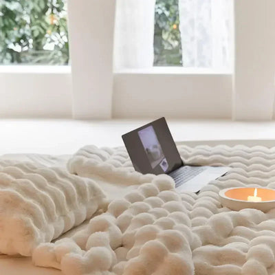 Lit douillet avec couverture texturée blanche, ordinateur portable ouvert et bougie allumée, ambiance chaleureuse et relaxante, intérieur moderne.