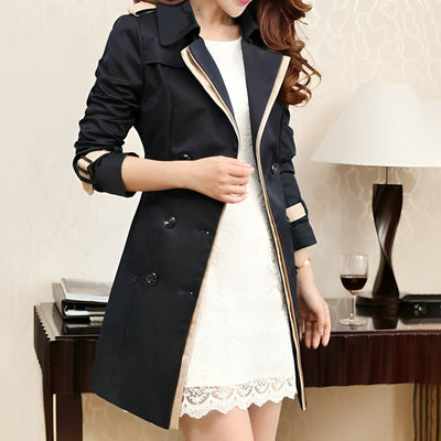 Femme portant un trench-coat noir élégant sur une robe blanche en dentelle, debout près d'une table avec une lampe et un verre de vin rouge.