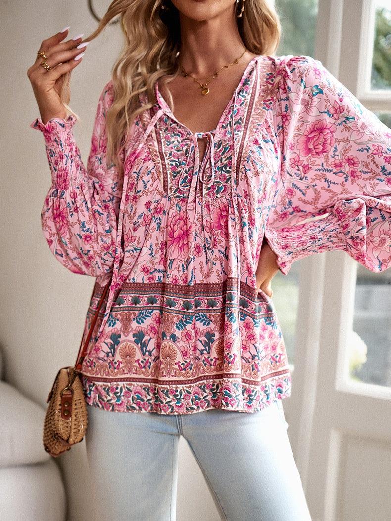 Femme portant un chemisier bohème rose à motifs floraux, manches longues bouffantes, avec un sac en osier, devant une fenêtre lumineuse.
