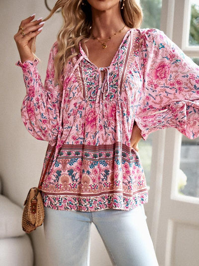 Femme portant un chemisier bohème rose à motifs floraux, manches longues bouffantes, avec un sac en osier, devant une fenêtre lumineuse.