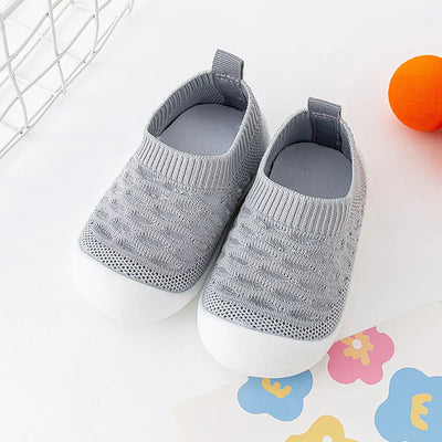 Chaussures bébé grises en tricot, semelles blanches antidérapantes, posées sur fond blanc avec jouet orange et papier fleuri coloré.