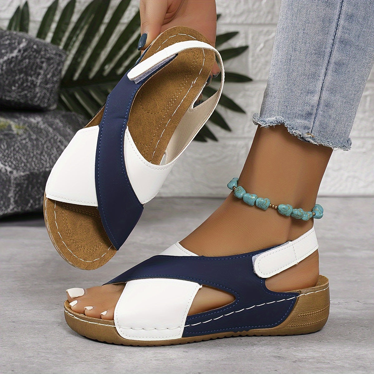 Sandales compensées bleu marine et blanches pour femme, portées avec un jean et un bracelet turquoise, sur fond de mur texturé et plantes.