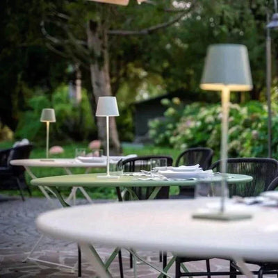 Tables de jardin élégantes avec lampes modernes, nappes blanches, verres et serviettes, entourées de verdure luxuriante. Ambiance extérieure paisible.