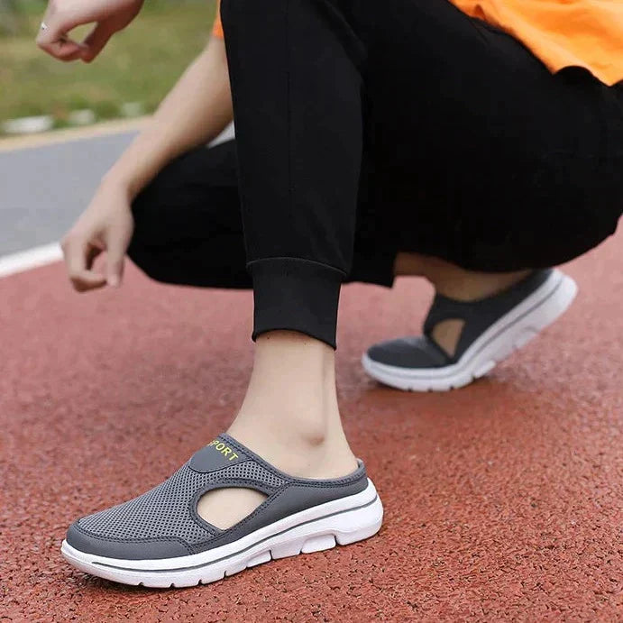 Personne portant des chaussures de sport grises sans lacets sur une piste rouge. Chaussures confortables, design respirant, semelle blanche.