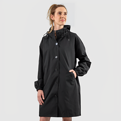 Femme portant un imperméable noir élégant avec capuche, boutons et poches, sur fond gris. Mode automne, vêtement de pluie tendance.