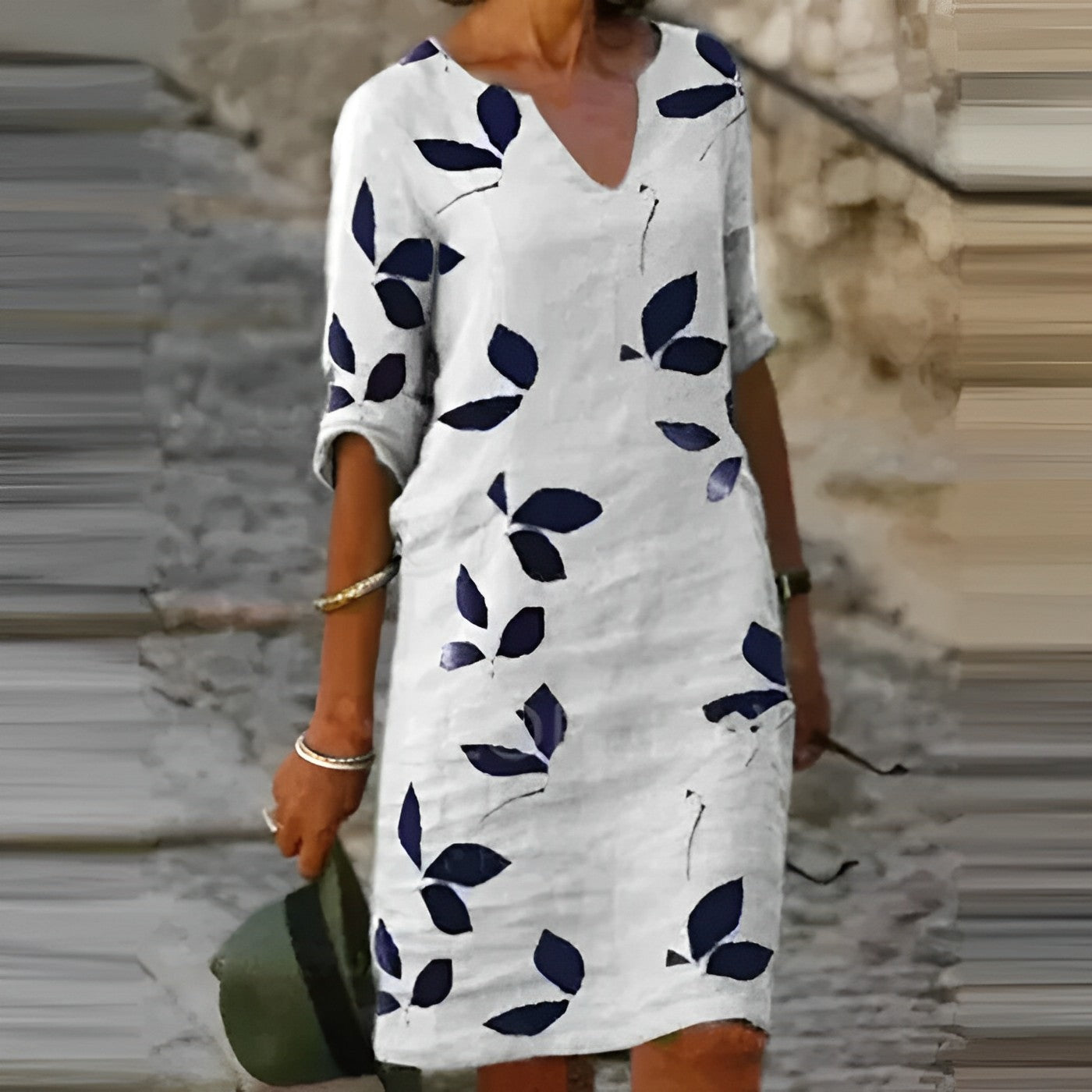 Robe blanche à motifs de feuilles bleues, manches trois-quarts, portée par une femme tenant un chapeau vert, sur fond de rue pavée.
