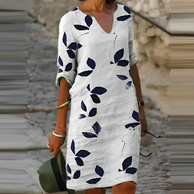 Robe blanche à motifs de feuilles bleues, manches trois-quarts, portée par une femme tenant un chapeau vert, sur fond de rue pavée.
