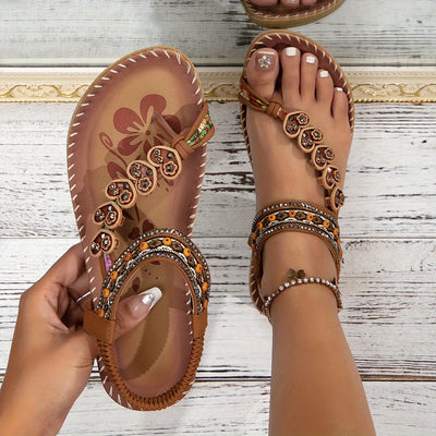 Sandales bohèmes marron avec motifs floraux et perles, portées sur pieds féminins avec pédicure blanche, sur fond de bois clair.