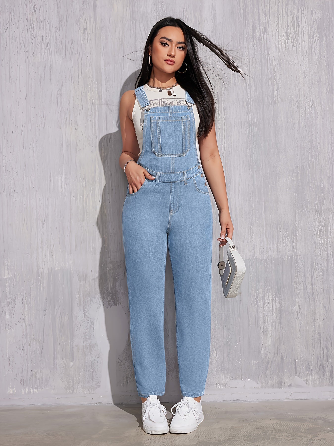 Femme portant une salopette en jean bleu clair, t-shirt blanc, baskets blanches, tenant un petit sac à main argenté, posant contre un mur gris.
