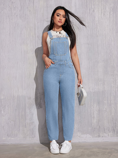 Femme portant une salopette en jean bleu clair, t-shirt blanc, baskets blanches, tenant un petit sac à main argenté, posant contre un mur gris.