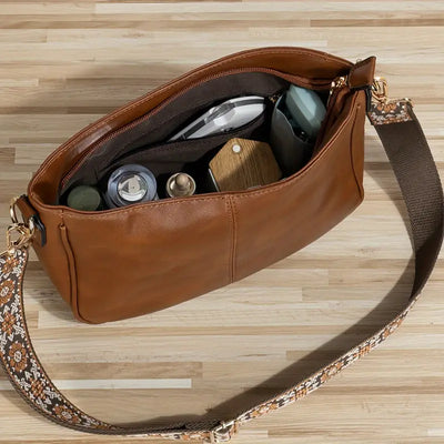 Sac à bandoulière en cuir marron ouvert sur une table en bois, contenant des articles de toilette et accessoires, avec sangle à motif floral.