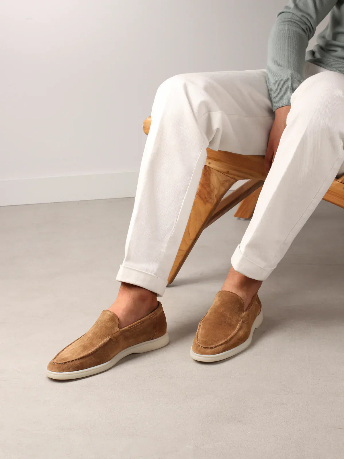 Homme assis portant des mocassins en daim marron clair, pantalon blanc et pull vert, sur un banc en bois. Mode masculine élégante et décontractée.