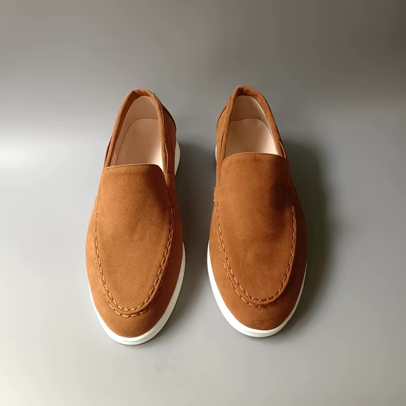 Paire de mocassins marron en daim sur fond gris, chaussures élégantes pour homme, semelle blanche, style décontracté et moderne.