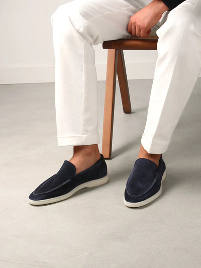 Homme portant des mocassins en daim bleu marine avec semelles blanches, assis sur une chaise, pantalon blanc, sol gris. Mode masculine élégante.