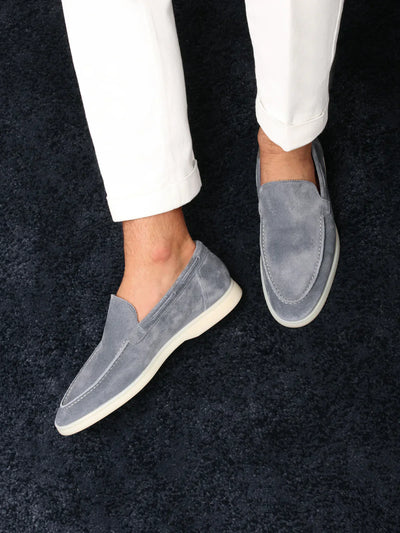 Chaussures mocassins en daim gris pour homme, portées avec un pantalon blanc sur un tapis bleu foncé. Style élégant et décontracté.