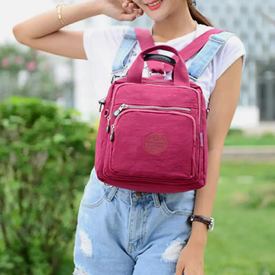 Femme portant un sac à dos rose en toile avec poches zippées, tenue décontractée en jean, extérieur verdoyant. Mode féminine, accessoires tendance.