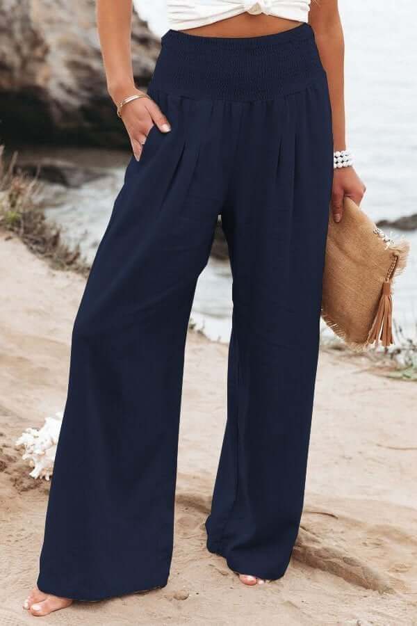 Pantalon large bleu marine femme, plage en arrière-plan, tenue estivale décontractée, accessoire sac en paille, mode bord de mer.