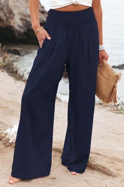 Pantalon large bleu marine femme, plage en arrière-plan, tenue estivale décontractée, accessoire sac en paille, mode bord de mer.