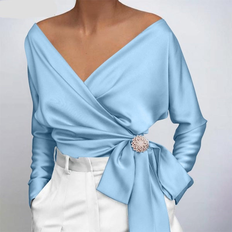 Blouse cache-cœur bleu clair élégante avec ceinture nouée, ornée d'une broche florale, portée avec un pantalon blanc taille haute. Mode féminine chic.