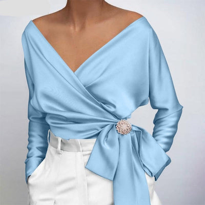 Blouse cache-cœur bleu clair élégante avec ceinture nouée, ornée d'une broche florale, portée avec un pantalon blanc taille haute. Mode féminine chic.