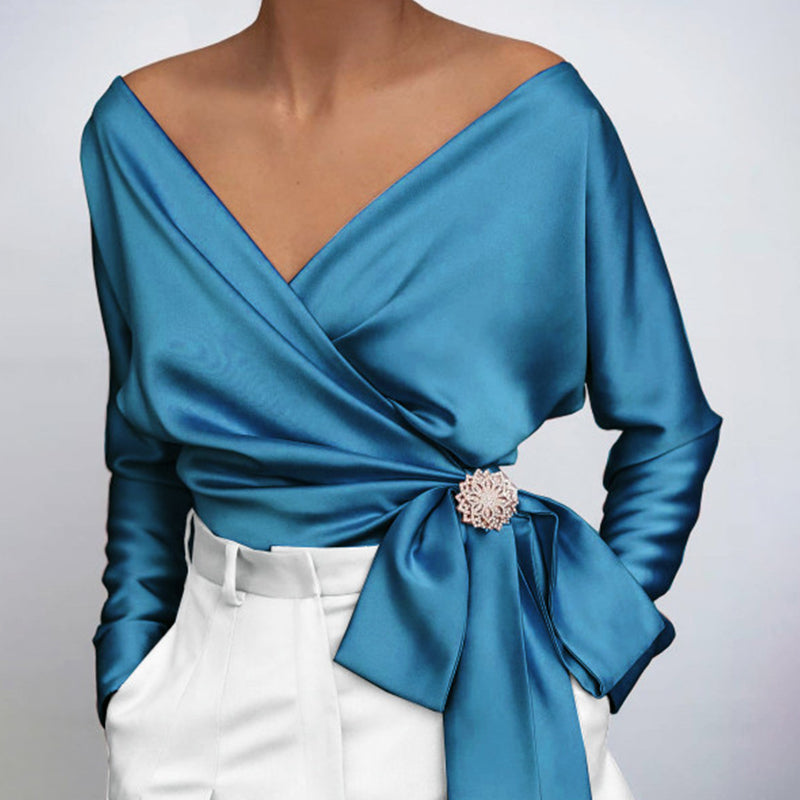 Blouse en satin bleu avec décolleté en V, manches longues, ceinture nouée et broche décorative, portée avec un pantalon blanc élégant.