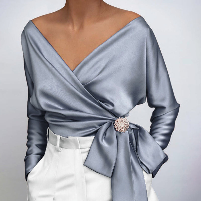 Blouse en satin gris perle avec décolleté en V, manches longues, nœud décoratif et broche florale, portée avec un pantalon blanc élégant.